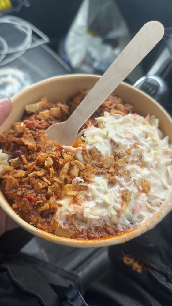 A hand holding a kraft bowl of jacket potato with cheese, sour cream and crispy onions, wooden fork in the bowl