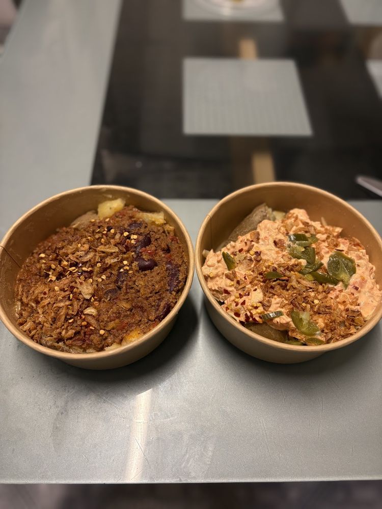 Two kraft bowls of jacket potato side by side on the steel counter, one chilli and crispy onion, one tuna mayo